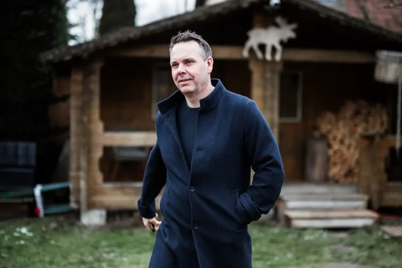 Thomas Kresse in einem dunklen Mantel gehend vor einer rustikalen Holzhütte – Bodenständigkeit und Tatkraft.